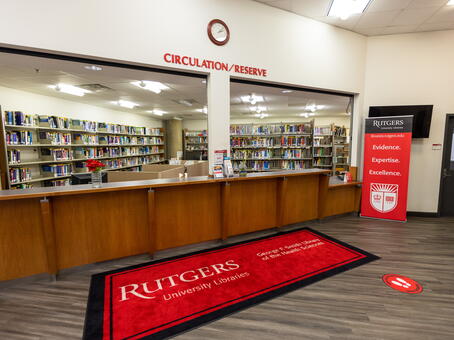 Image of the circulation and reserve desk at the Smith library, with no staff in the image, but stacks of books behind the counter. There is a large red mat in front of the counter that reads "Rutgers University Libraries" as well as lettering above the counter stating Circulation/Reserve