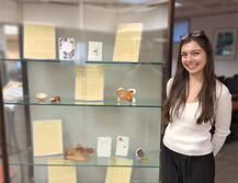 Image of student artist Sophia Westfall standing next to a display case filled with her work.